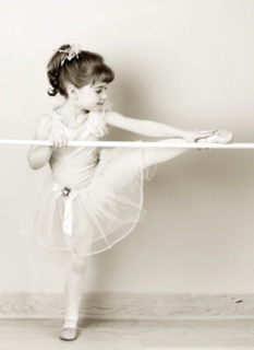 A girl wearing a ballet outfit stretches her arm to her foot which is perched on a ballet bar.