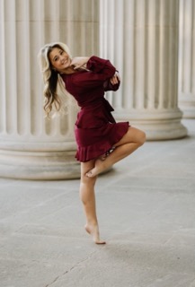 A woman in a red dress poses on tip-toe with one leg lifted to her knee. Her arms are folded so her hands are on her chin. She smiles at the camera.