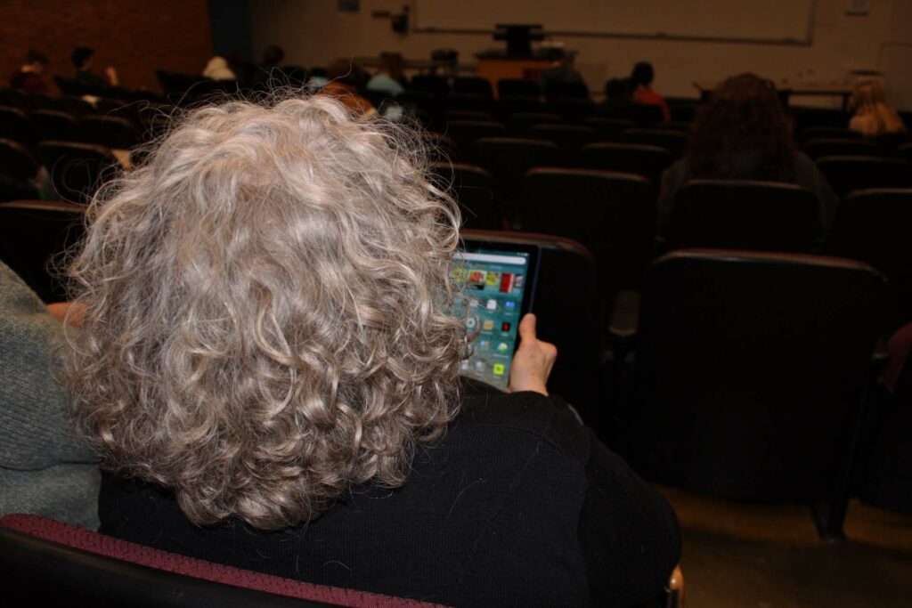 A woman sits at the back of a classroom facing toward the front of the room, she holds an iPad.