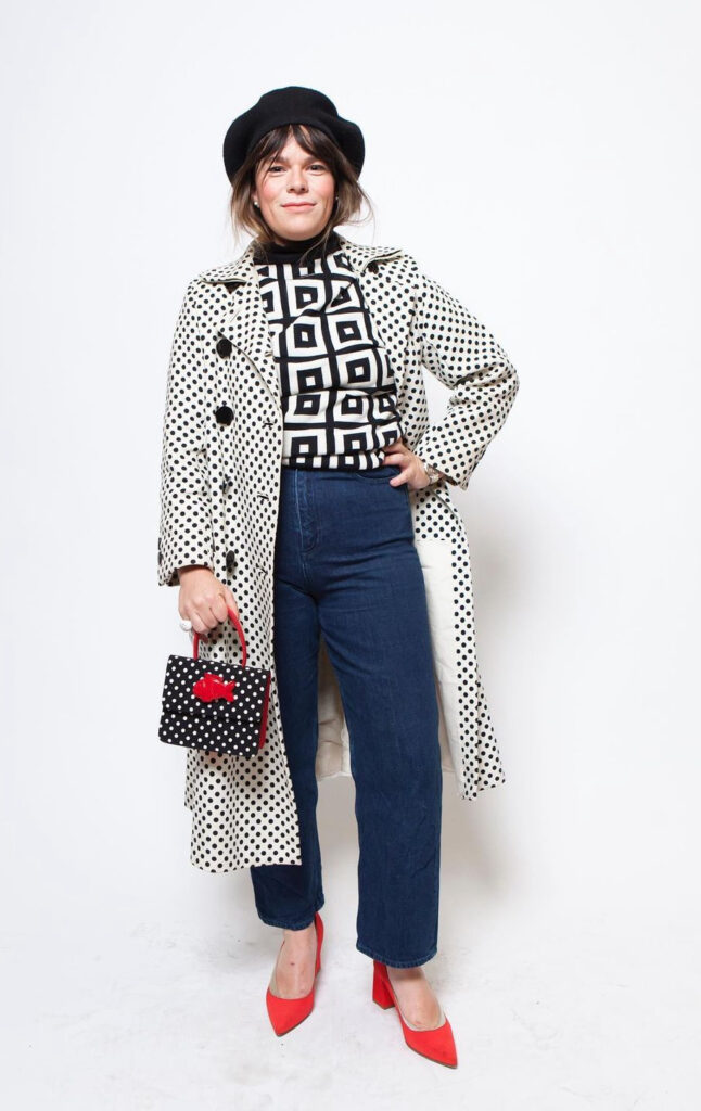Full body photo of a woman posing with her hand on her hip. She is wearing red heels, bluejeans, a black and white square patterned shirt, and a black and white polka dot trench coat. The outfit is accessorized with a black brim hat and a black and white polkadot purse with red accents.