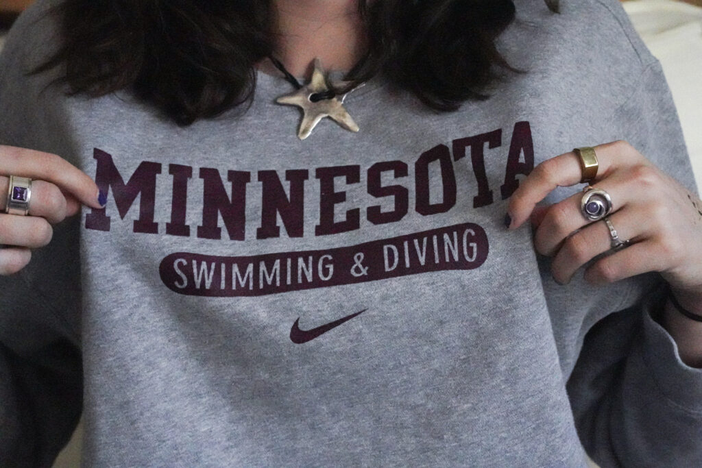 A person pulls at their sweatshirt, showing the logo. The shirt says Minnesota Swimming & Diving in maroon text.