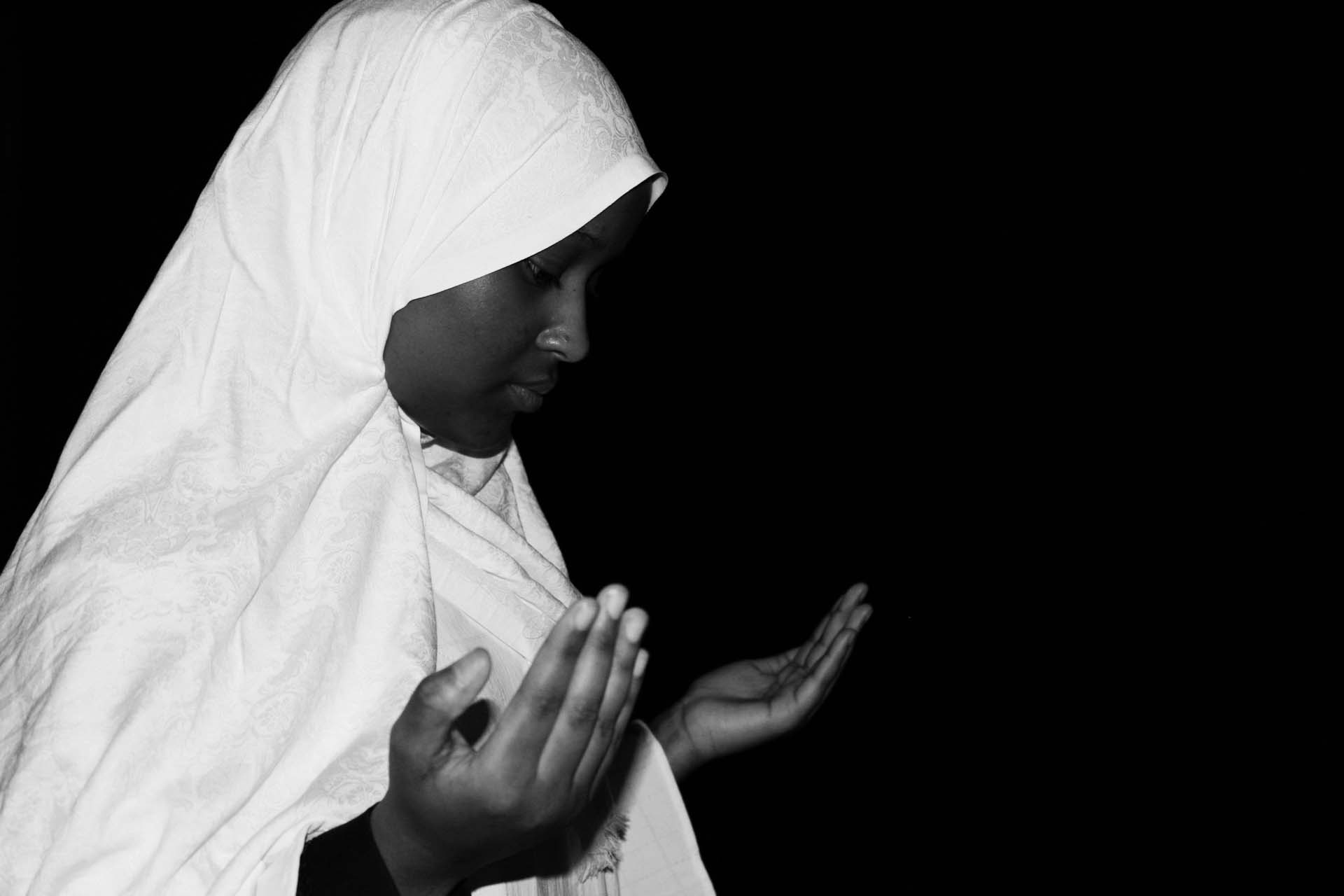 A woman poses with her hands up in prayer looking down, she wears a white cloth covering her from head to toe.