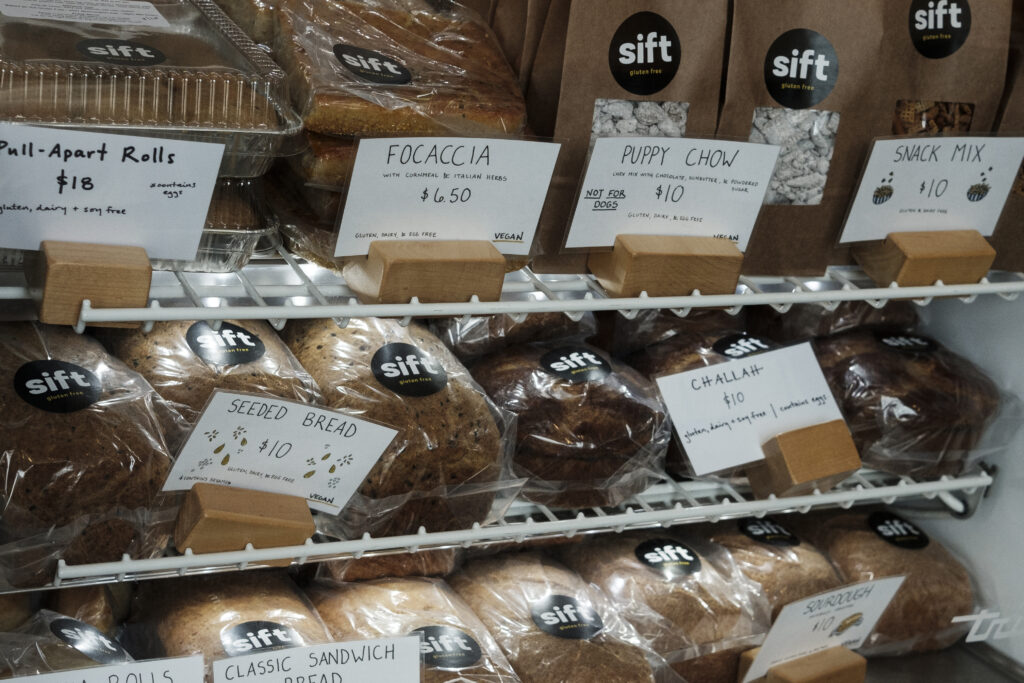 Multiple loafs of bread wrapped in plastic in a display case. In front of the bread is a sign that reads “Seeded Bread $10 / Gluten, Dairy, & Egg Free”