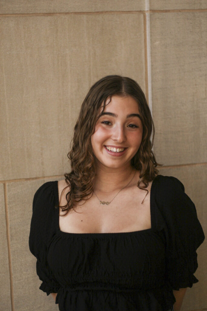 A girl looks at the viewer and smiles. She wears a necklace and black shirt.