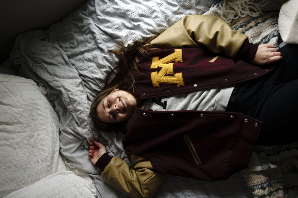 Arial view of a person lying on a gray bedspread wearing a University of Minnesota varsity letterman jacket. They are smiling at the camera.