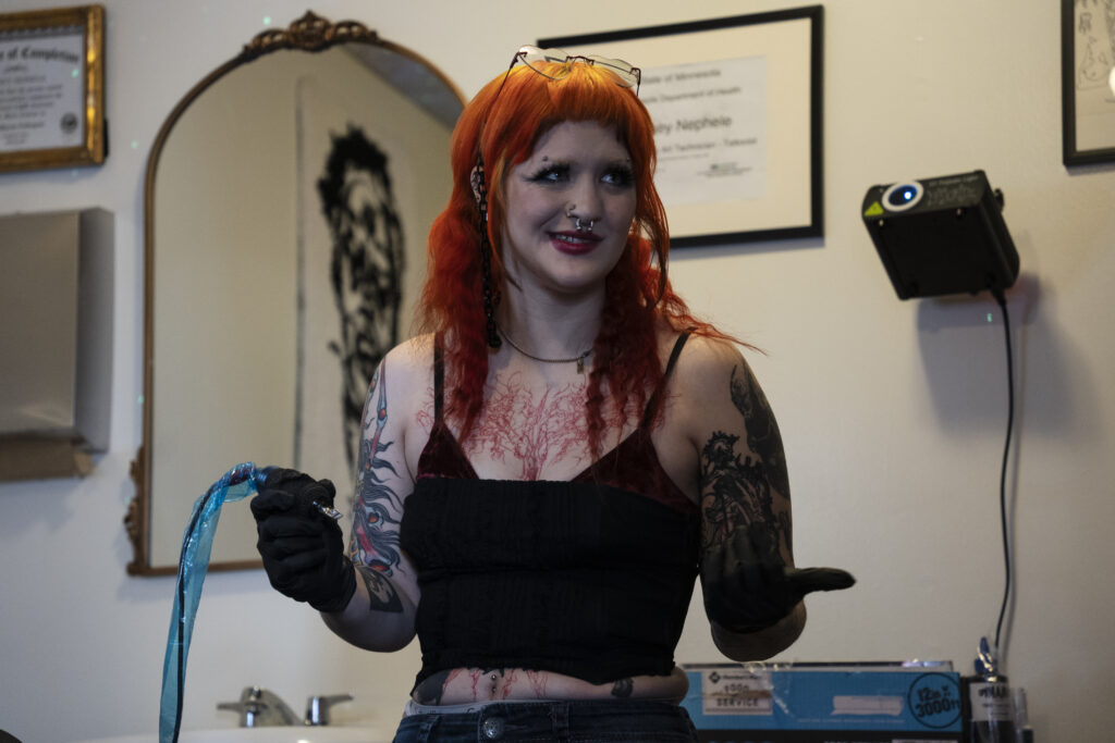 A woman stands in a room wearing black gloves and holds a tattoo gun in their right hand. They are looking to the right and are shrugging with a smirk.